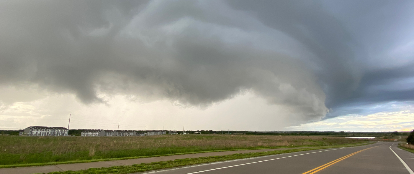 Large, dark storm clouds over a winding rural highway, with grassy fields and distant horizon beneath the approaching weather.